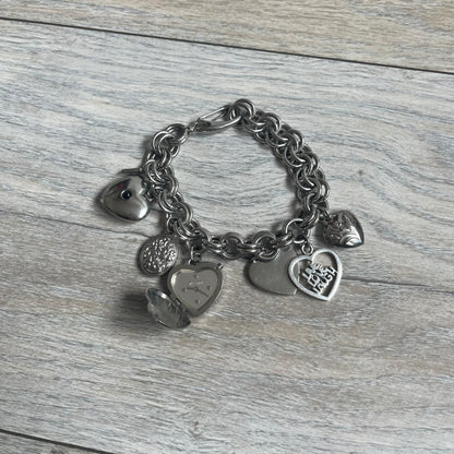 Moschino 2000s “Live Love Laugh” Charms Bracelet Watch is a stainless steel bracelet watch with a silver-tone finish. The bracelet is made of connected circular links and features multiple hanging heart-shaped charms. One charm displays “Live Love Laugh” text, while the main charm is a heart-shaped opening watch case with an “M” on the front. The watch dial is small and uses heart symbols in place of the main hour markers. All components are finished in silver tone.