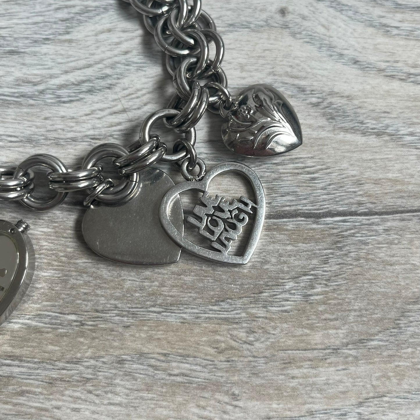 Moschino 2000s “Live Love Laugh” Charms Bracelet Watch is a stainless steel bracelet watch with a silver-tone finish. The bracelet is made of connected circular links and features multiple hanging heart-shaped charms. One charm displays “Live Love Laugh” text, while the main charm is a heart-shaped opening watch case with an “M” on the front. The watch dial is small and uses heart symbols in place of the main hour markers. All components are finished in silver tone.