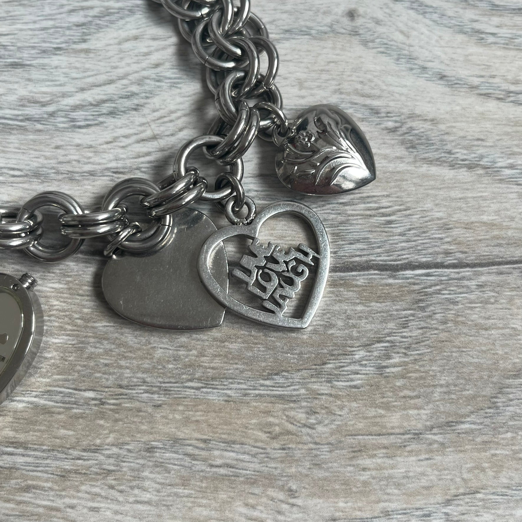 Moschino 2000s “Live Love Laugh” Charms Bracelet Watch is a stainless steel bracelet watch with a silver-tone finish. The bracelet is made of connected circular links and features multiple hanging heart-shaped charms. One charm displays “Live Love Laugh” text, while the main charm is a heart-shaped opening watch case with an “M” on the front. The watch dial is small and uses heart symbols in place of the main hour markers. All components are finished in silver tone.