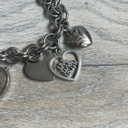 Moschino 2000s “Live Love Laugh” Charms Bracelet Watch is a stainless steel bracelet watch with a silver-tone finish. The bracelet is made of connected circular links and features multiple hanging heart-shaped charms. One charm displays “Live Love Laugh” text, while the main charm is a heart-shaped opening watch case with an “M” on the front. The watch dial is small and uses heart symbols in place of the main hour markers. All components are finished in silver tone.
