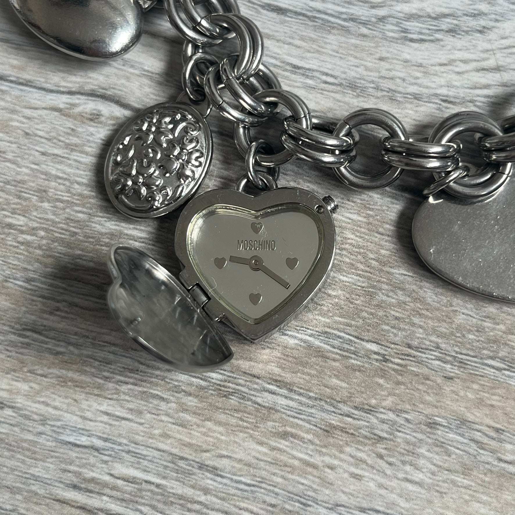 Moschino 2000s “Live Love Laugh” Charms Bracelet Watch is a stainless steel bracelet watch with a silver-tone finish. The bracelet is made of connected circular links and features multiple hanging heart-shaped charms. One charm displays “Live Love Laugh” text, while the main charm is a heart-shaped opening watch case with an “M” on the front. The watch dial is small and uses heart symbols in place of the main hour markers. All components are finished in silver tone.
