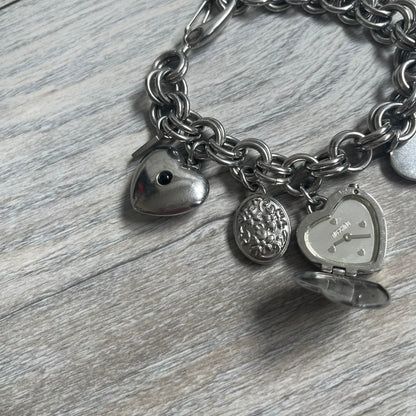 Moschino 2000s “Live Love Laugh” Charms Bracelet Watch is a stainless steel bracelet watch with a silver-tone finish. The bracelet is made of connected circular links and features multiple hanging heart-shaped charms. One charm displays “Live Love Laugh” text, while the main charm is a heart-shaped opening watch case with an “M” on the front. The watch dial is small and uses heart symbols in place of the main hour markers. All components are finished in silver tone.