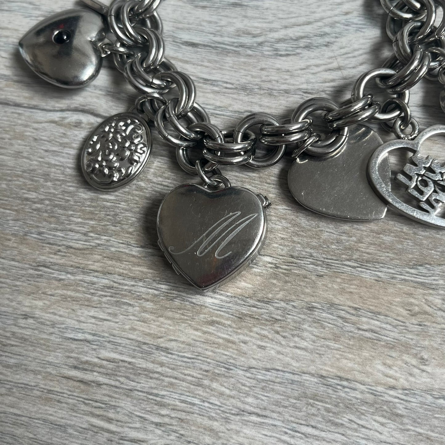 Moschino 2000s “Live Love Laugh” Charms Bracelet Watch is a stainless steel bracelet watch with a silver-tone finish. The bracelet is made of connected circular links and features multiple hanging heart-shaped charms. One charm displays “Live Love Laugh” text, while the main charm is a heart-shaped opening watch case with an “M” on the front. The watch dial is small and uses heart symbols in place of the main hour markers. All components are finished in silver tone.