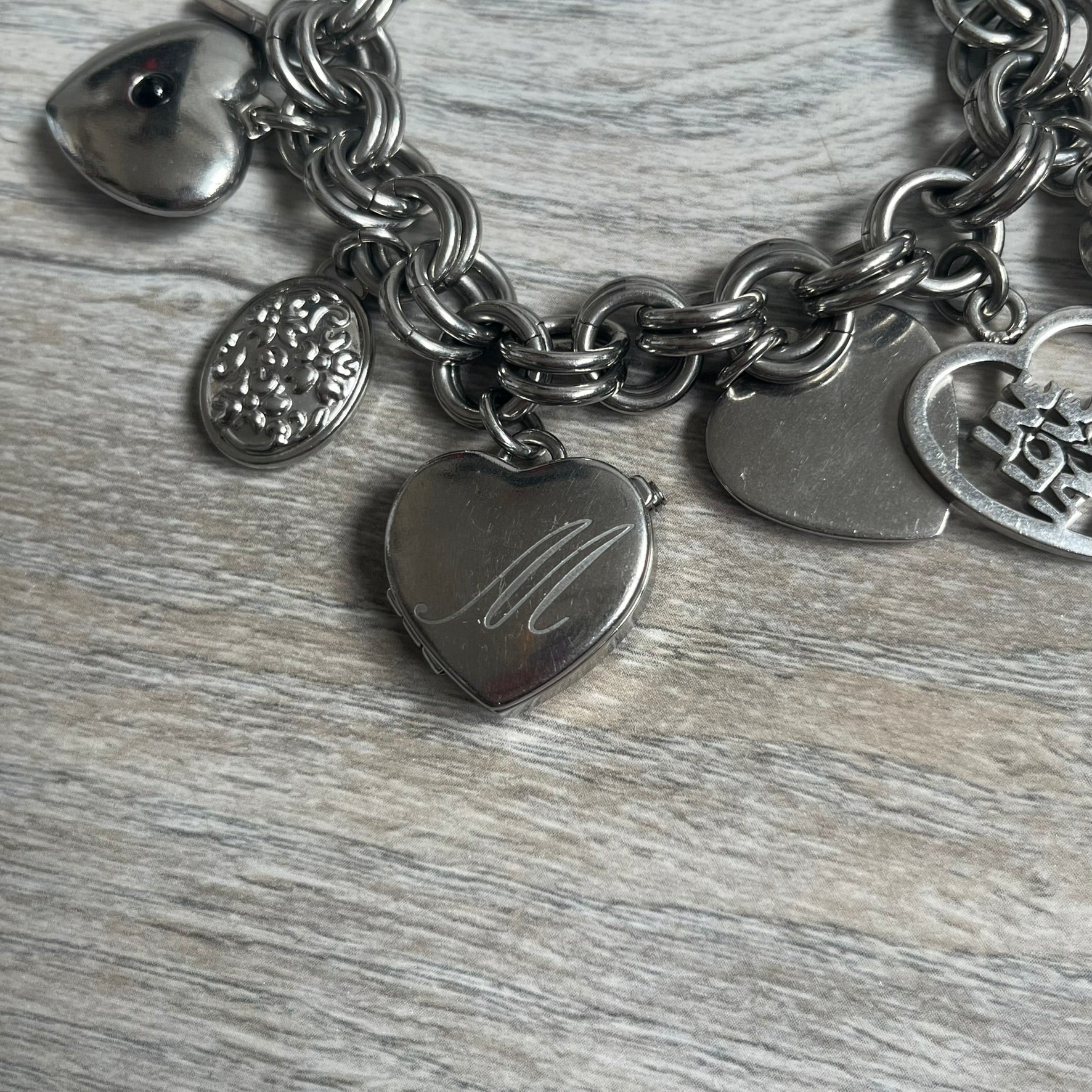 Moschino 2000s “Live Love Laugh” Charms Bracelet Watch is a stainless steel bracelet watch with a silver-tone finish. The bracelet is made of connected circular links and features multiple hanging heart-shaped charms. One charm displays “Live Love Laugh” text, while the main charm is a heart-shaped opening watch case with an “M” on the front. The watch dial is small and uses heart symbols in place of the main hour markers. All components are finished in silver tone.