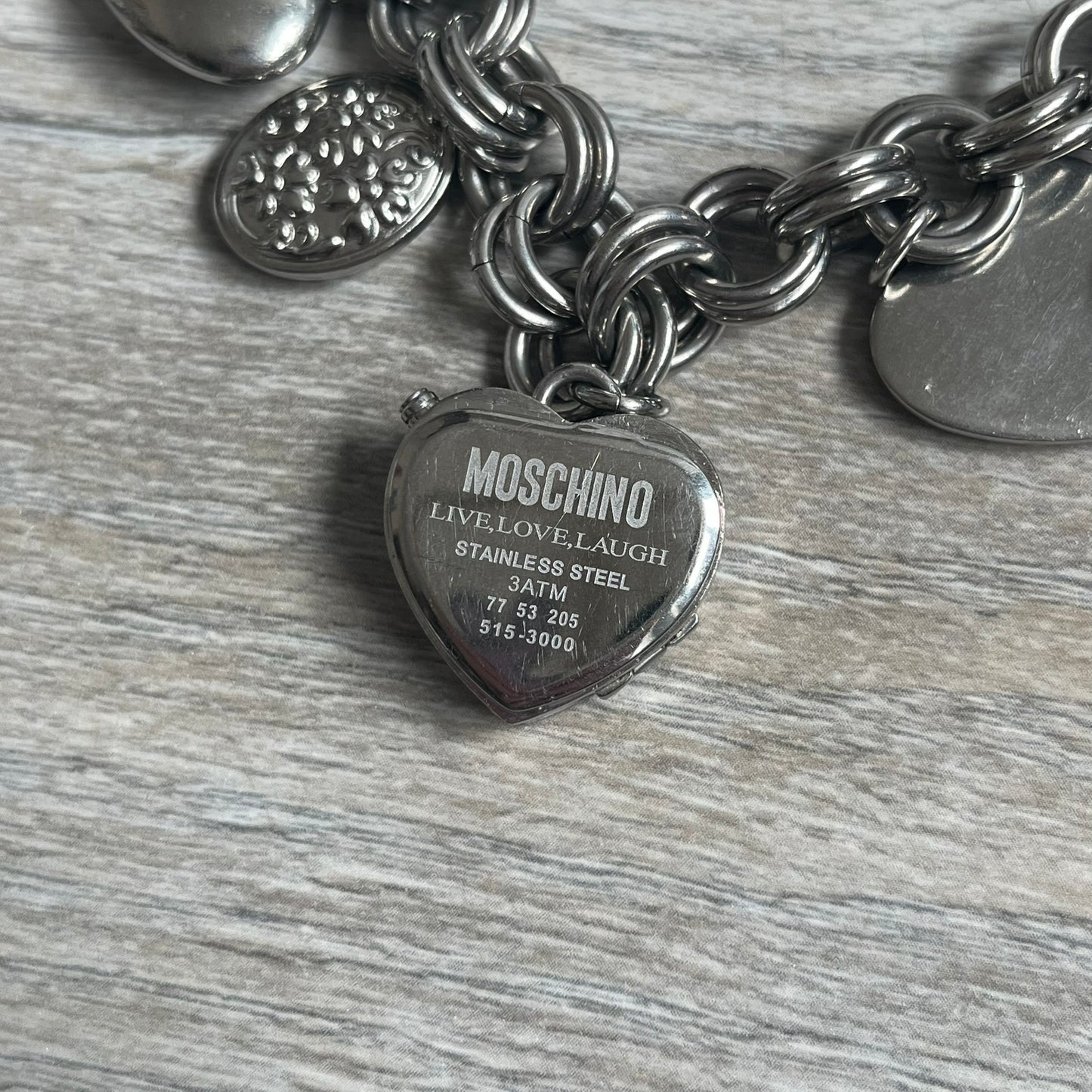 Moschino 2000s “Live Love Laugh” Charms Bracelet Watch is a stainless steel bracelet watch with a silver-tone finish. The bracelet is made of connected circular links and features multiple hanging heart-shaped charms. One charm displays “Live Love Laugh” text, while the main charm is a heart-shaped opening watch case with an “M” on the front. The watch dial is small and uses heart symbols in place of the main hour markers. All components are finished in silver tone.