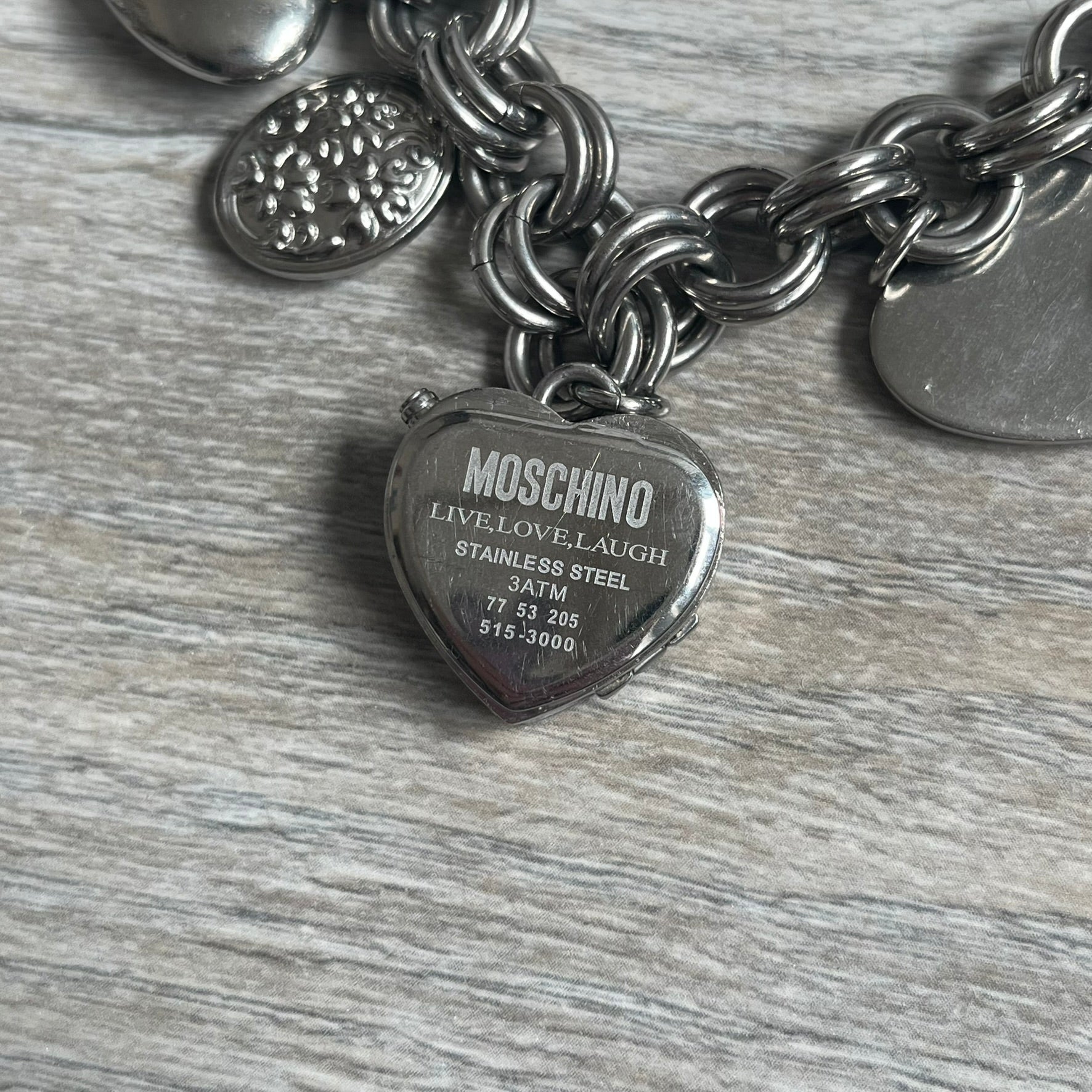 Moschino 2000s “Live Love Laugh” Charms Bracelet Watch is a stainless steel bracelet watch with a silver-tone finish. The bracelet is made of connected circular links and features multiple hanging heart-shaped charms. One charm displays “Live Love Laugh” text, while the main charm is a heart-shaped opening watch case with an “M” on the front. The watch dial is small and uses heart symbols in place of the main hour markers. All components are finished in silver tone.