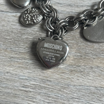 Moschino 2000s “Live Love Laugh” Charms Bracelet Watch is a stainless steel bracelet watch with a silver-tone finish. The bracelet is made of connected circular links and features multiple hanging heart-shaped charms. One charm displays “Live Love Laugh” text, while the main charm is a heart-shaped opening watch case with an “M” on the front. The watch dial is small and uses heart symbols in place of the main hour markers. All components are finished in silver tone.
