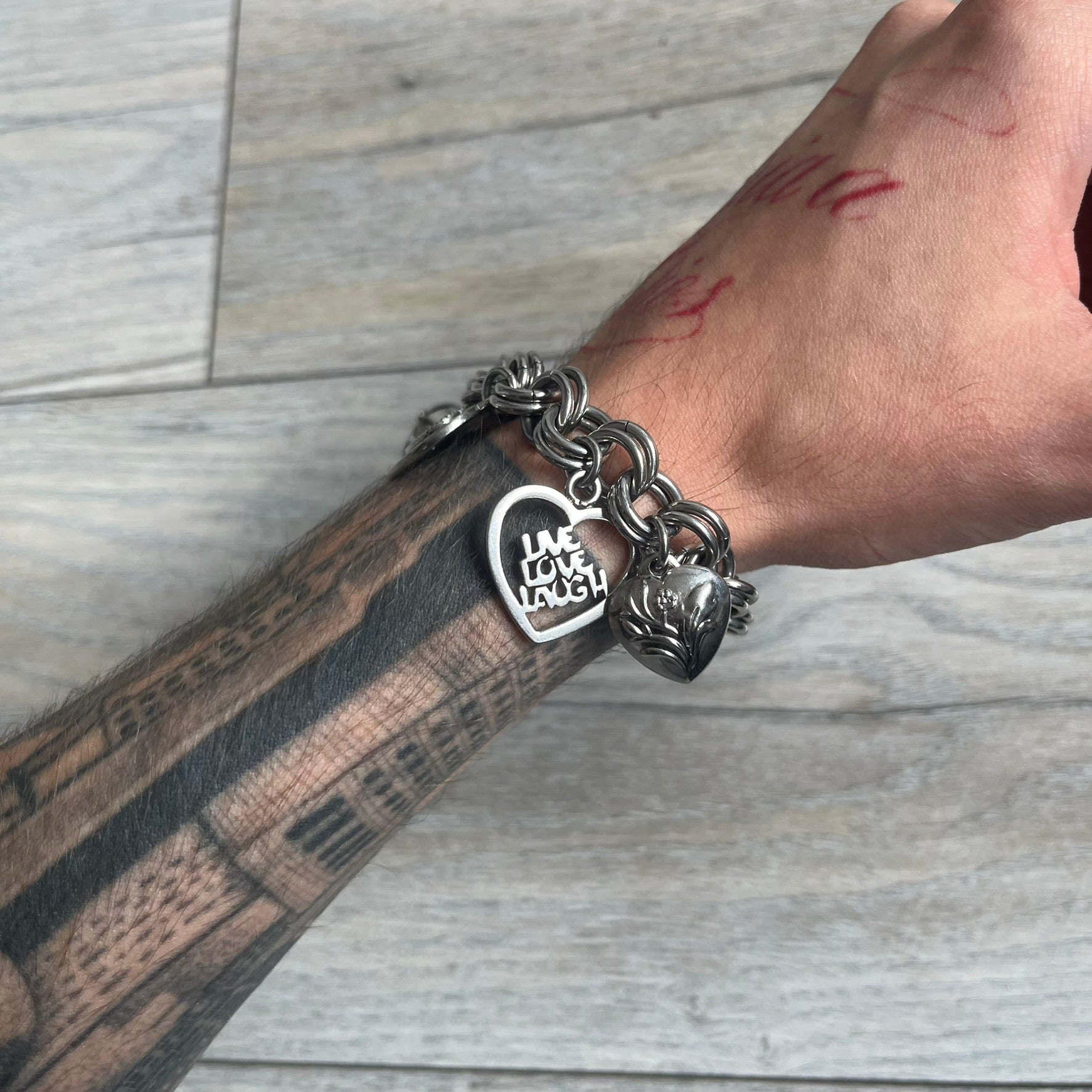 Moschino 2000s “Live Love Laugh” Charms Bracelet Watch is a stainless steel bracelet watch with a silver-tone finish. The bracelet is made of connected circular links and features multiple hanging heart-shaped charms. One charm displays “Live Love Laugh” text, while the main charm is a heart-shaped opening watch case with an “M” on the front. The watch dial is small and uses heart symbols in place of the main hour markers. All components are finished in silver tone.