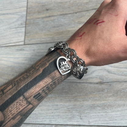 Moschino 2000s “Live Love Laugh” Charms Bracelet Watch is a stainless steel bracelet watch with a silver-tone finish. The bracelet is made of connected circular links and features multiple hanging heart-shaped charms. One charm displays “Live Love Laugh” text, while the main charm is a heart-shaped opening watch case with an “M” on the front. The watch dial is small and uses heart symbols in place of the main hour markers. All components are finished in silver tone.
