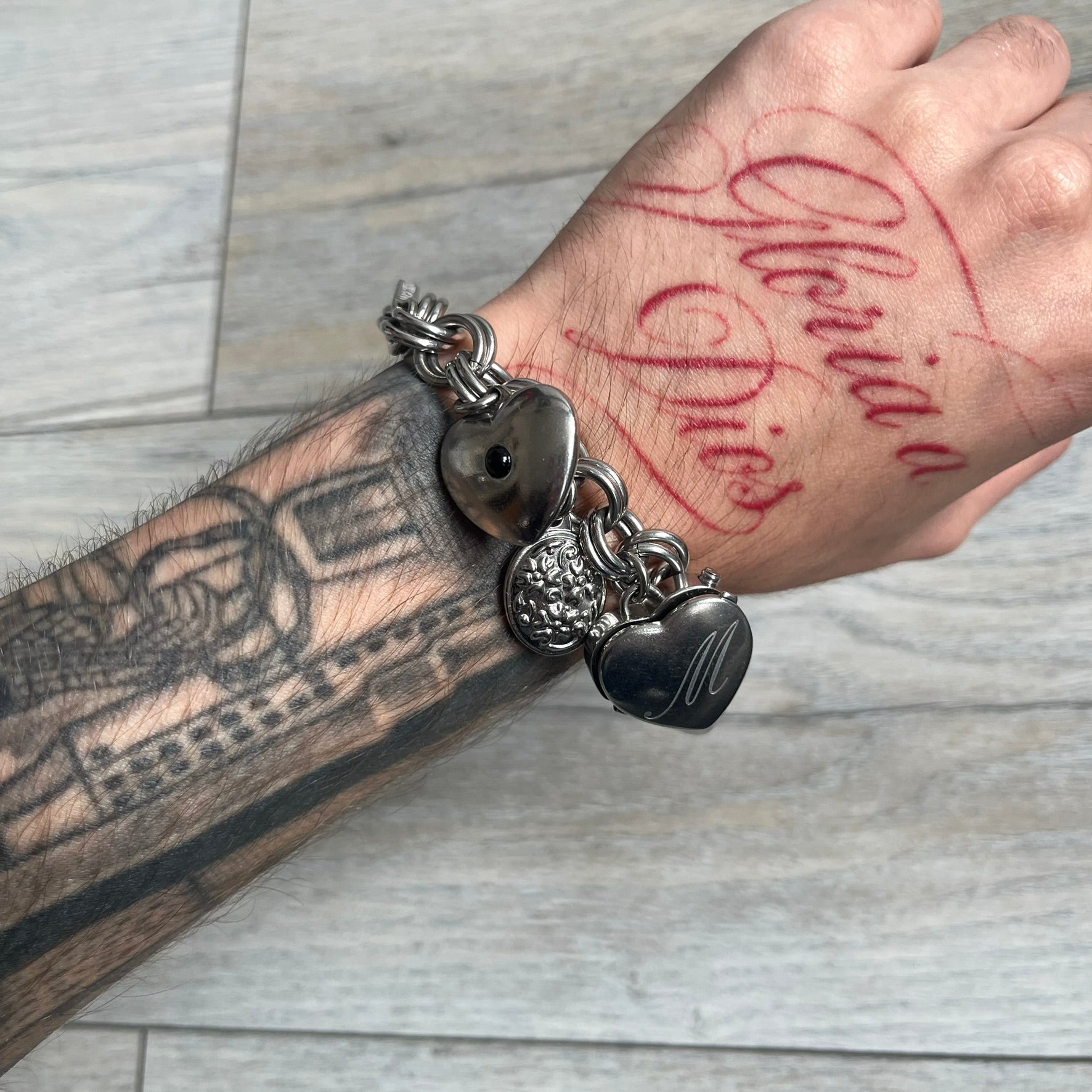 Moschino 2000s “Live Love Laugh” Charms Bracelet Watch is a stainless steel bracelet watch with a silver-tone finish. The bracelet is made of connected circular links and features multiple hanging heart-shaped charms. One charm displays “Live Love Laugh” text, while the main charm is a heart-shaped opening watch case with an “M” on the front. The watch dial is small and uses heart symbols in place of the main hour markers. All components are finished in silver tone.