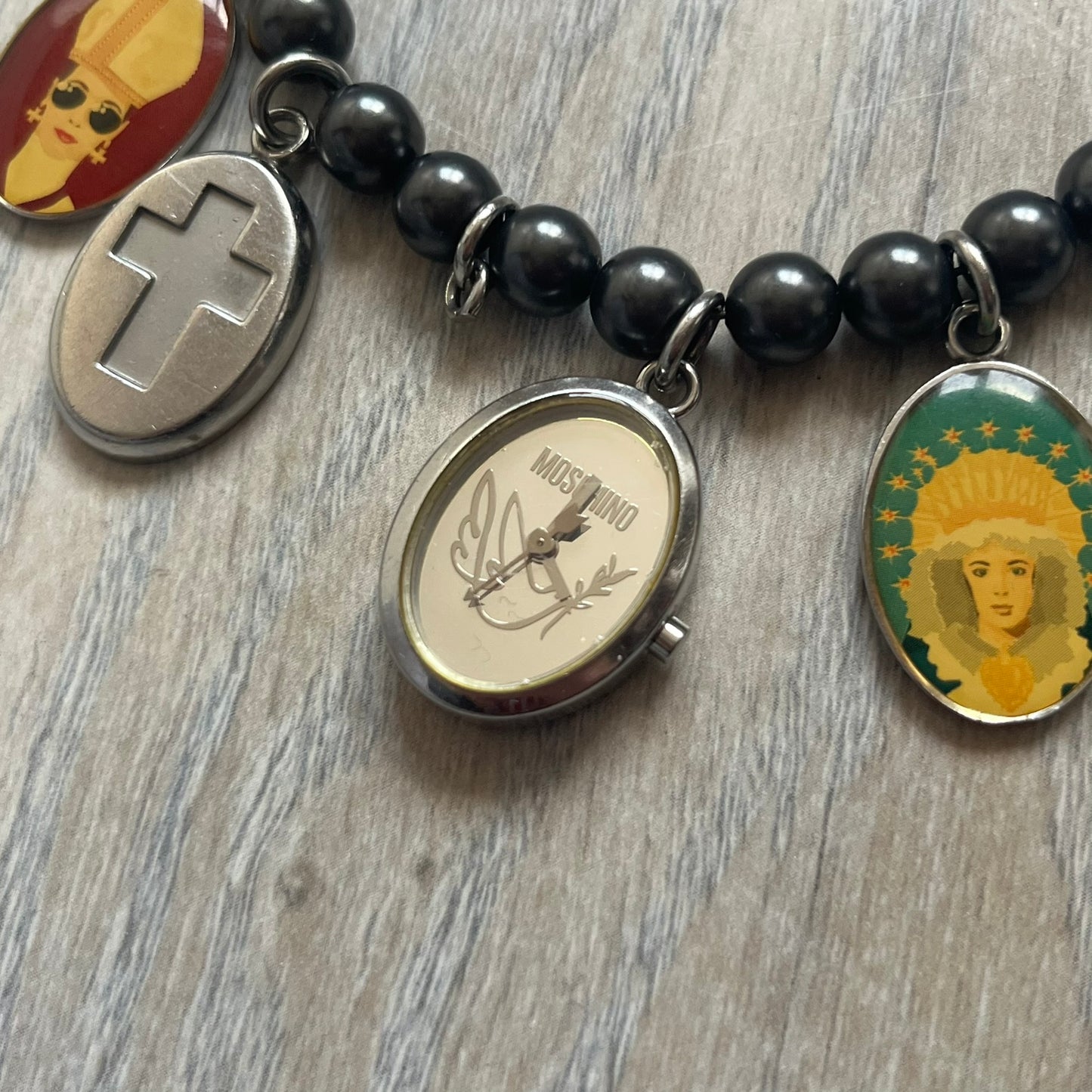 Moschino 2000’s “Time 4 Holy” Pendants Bracelet Watch is a bracelet watch from Moschino featuring a beaded construction with black pearly beads and multiple hanging religious-themed charms. The bracelet includes Madonna and holy figure pendants, with one charm functioning as a small hour-and-minute watch. Made in Italy in the early 2000s, the piece uses a pendant-style layout combining jewelry elements with a functional watch.