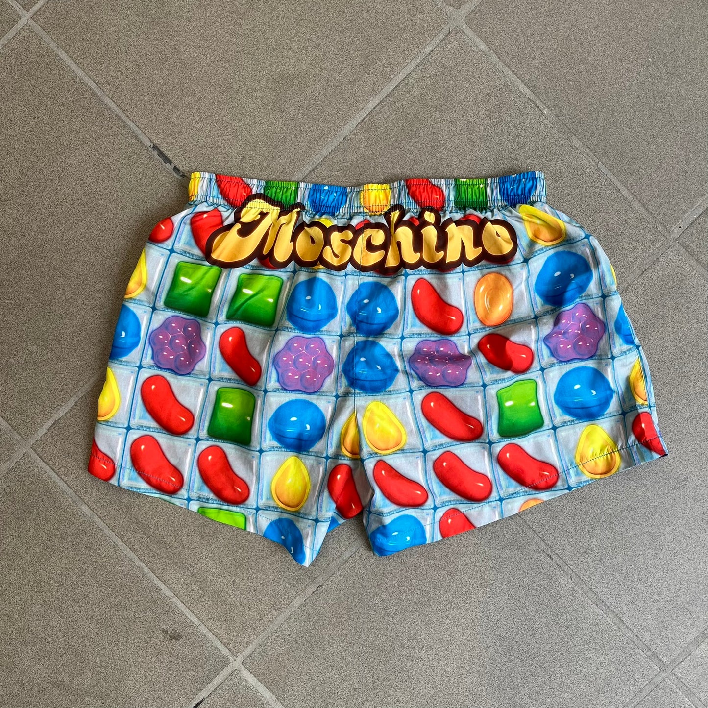 Moschino Candy Crush 2017 Swimming Shorts feature an all-over print inspired by the Candy Crush game board, with colorful candy graphics covering the fabric. The back is detailed with a large Moschino logo in a gold Candy Crush-style font. The shorts include red drawstrings with Moschino-branded metal aglets, a mesh inner lining, and are made in Italy.
