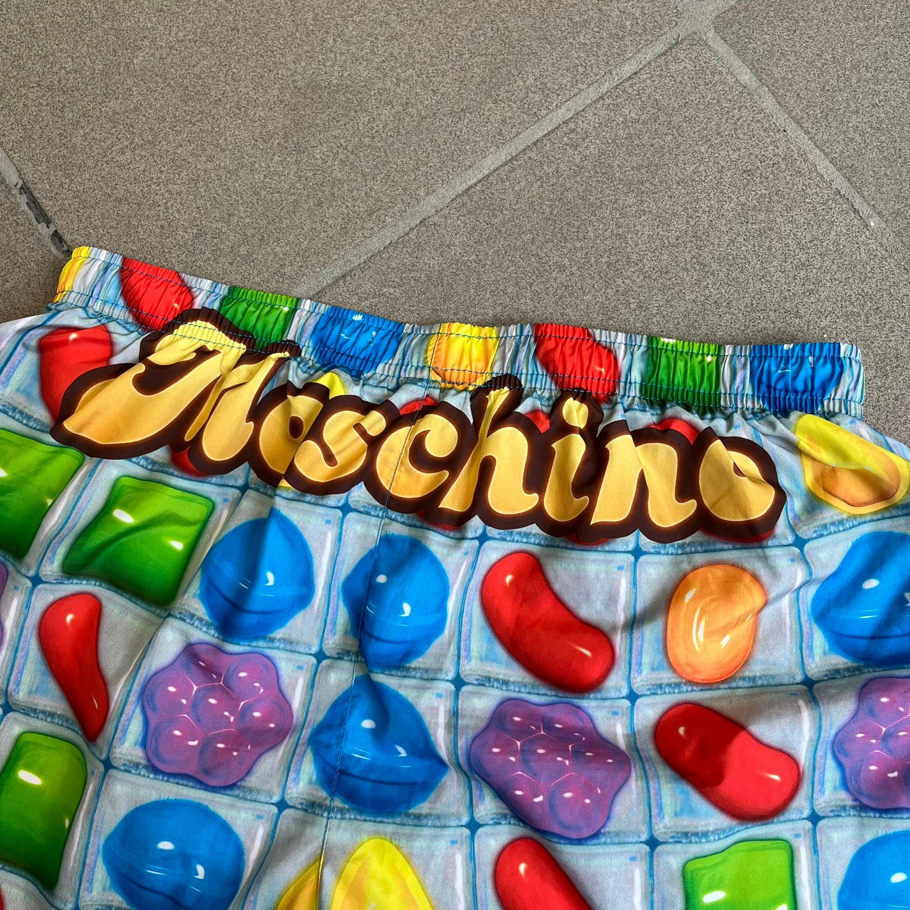 Moschino Candy Crush 2017 Swimming Shorts feature an all-over print inspired by the Candy Crush game board, with colorful candy graphics covering the fabric. The back is detailed with a large Moschino logo in a gold Candy Crush-style font. The shorts include red drawstrings with Moschino-branded metal aglets, a mesh inner lining, and are made in Italy.
