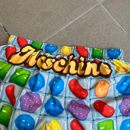 Moschino Candy Crush 2017 Swimming Shorts feature an all-over print inspired by the Candy Crush game board, with colorful candy graphics covering the fabric. The back is detailed with a large Moschino logo in a gold Candy Crush-style font. The shorts include red drawstrings with Moschino-branded metal aglets, a mesh inner lining, and are made in Italy.
