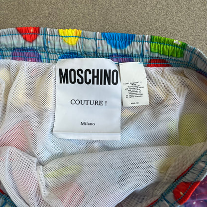 Moschino Candy Crush 2017 Swimming Shorts feature an all-over print inspired by the Candy Crush game board, with colorful candy graphics covering the fabric. The back is detailed with a large Moschino logo in a gold Candy Crush-style font. The shorts include red drawstrings with Moschino-branded plastic aglets, a mesh inner lining, and are made in Italy.
