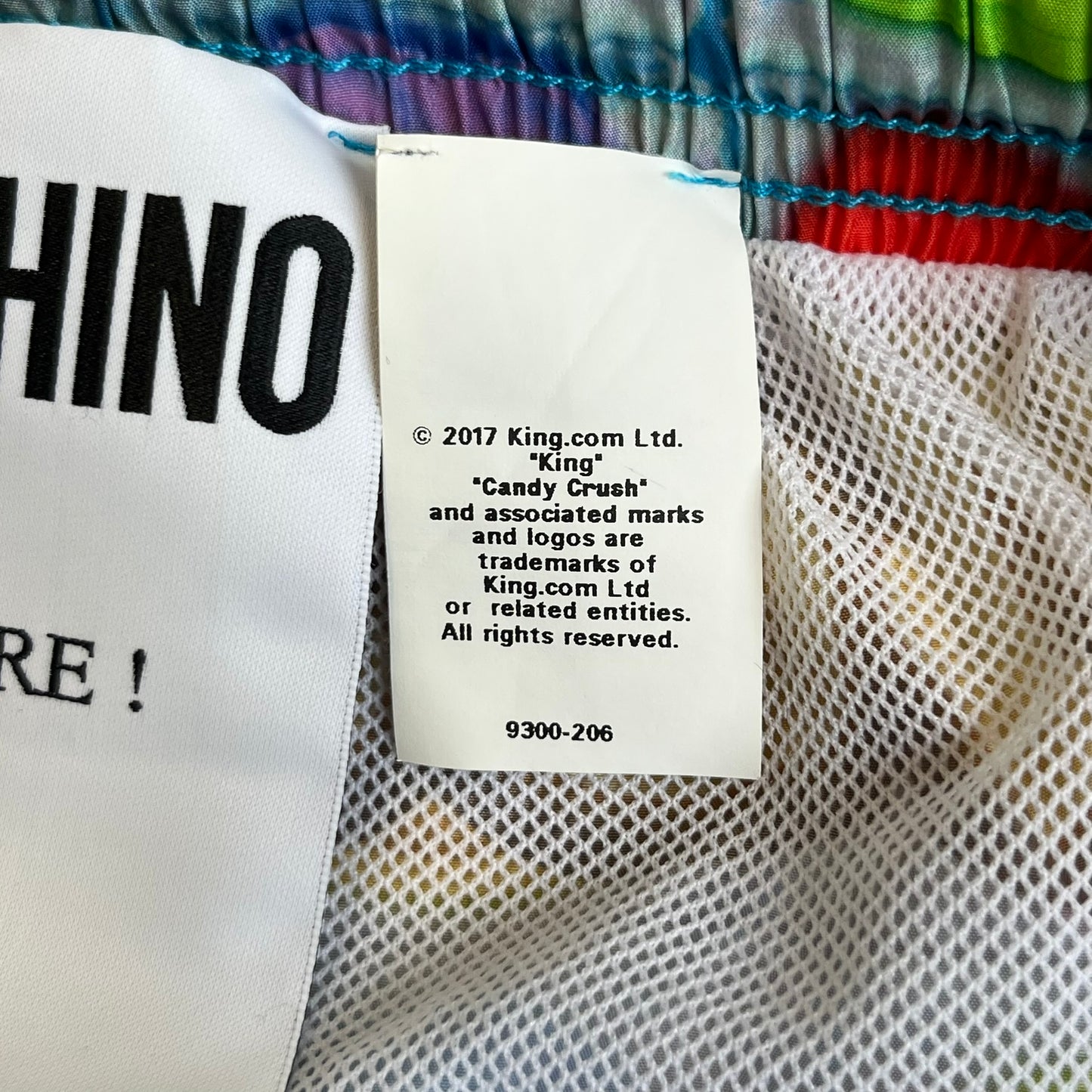 Moschino Candy Crush 2017 Swimming Shorts feature an all-over print inspired by the Candy Crush game board, with colorful candy graphics covering the fabric. The back is detailed with a large Moschino logo in a gold Candy Crush-style font. The shorts include red drawstrings with Moschino-branded plastic aglets, a mesh inner lining, and are made in Italy.

