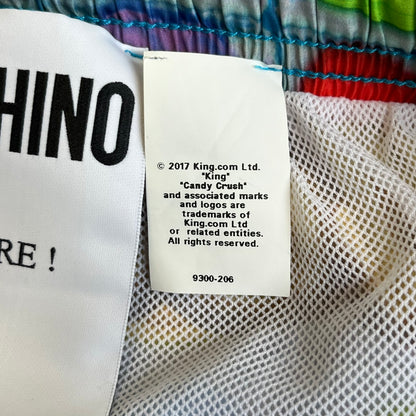 Moschino Candy Crush 2017 Swimming Shorts feature an all-over print inspired by the Candy Crush game board, with colorful candy graphics covering the fabric. The back is detailed with a large Moschino logo in a gold Candy Crush-style font. The shorts include red drawstrings with Moschino-branded plastic aglets, a mesh inner lining, and are made in Italy.

