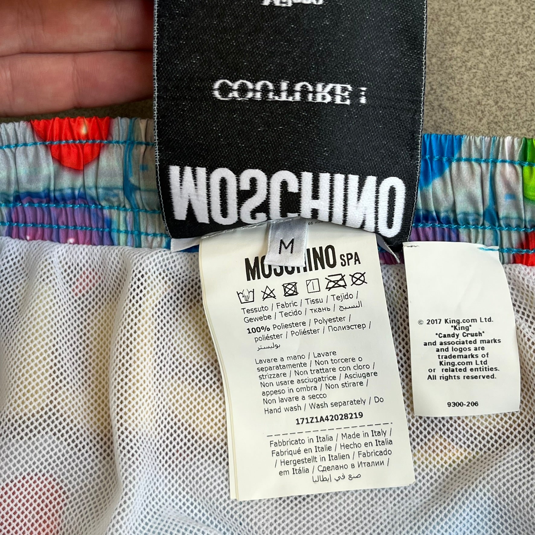 Moschino Candy Crush 2017 Swimming Shorts feature an all-over print inspired by the Candy Crush game board, with colorful candy graphics covering the fabric. The back is detailed with a large Moschino logo in a gold Candy Crush-style font. The shorts include red drawstrings with Moschino-branded plastic aglets, a mesh inner lining, and are made in Italy.
