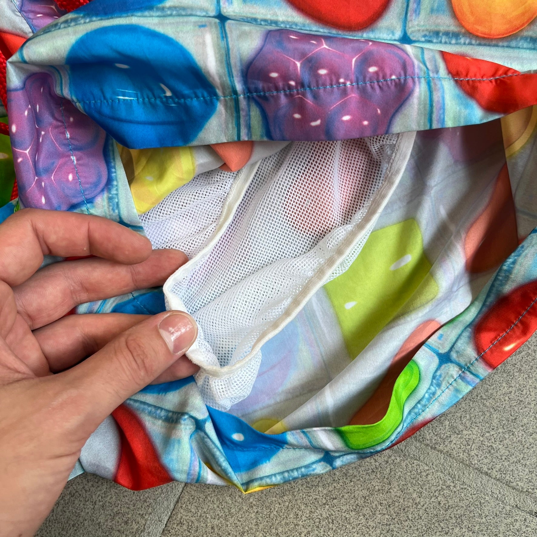 Moschino Candy Crush 2017 Swimming Shorts feature an all-over print inspired by the Candy Crush game board, with colorful candy graphics covering the fabric. The back is detailed with a large Moschino logo in a gold Candy Crush-style font. The shorts include red drawstrings with Moschino-branded plastic aglets, a mesh inner lining, and are made in Italy.
