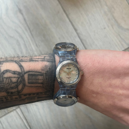 Moschino “Don’t Be 2 Late” Clear Blue 5 Dial Analog Watch in a light blue colorway, featuring an extended plastic strap and case construction. The design includes five separate dials with individual movements: four pearlescent faces in varying tones and one fully transparent dial, each with different markers including Roman numerals, Arabic numerals, or crystal hour indices. Silver-tone metal bezel rings and crowns (smooth, ridged, and crystal-set variations) frame the dials, while stainless steel casebacks