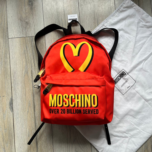 Moschino FW14 McDonald’s Fast Food Backpack is a red nylon backpack from the Autumn/Winter 2014 collection, designed by Jeremy Scott for his debut Moschino lineup. The front features yellow curved arches inspired by McDonald’s branding, with a front zip pocket printed with yellow Moschino text and black “Over 20 Billion Served” slogan. The bag includes gold-tone zippers with black leather pulls, black straps with Moschino logos, a branded interior lining, and comes with a Moschino dust bag. Made in Italy.