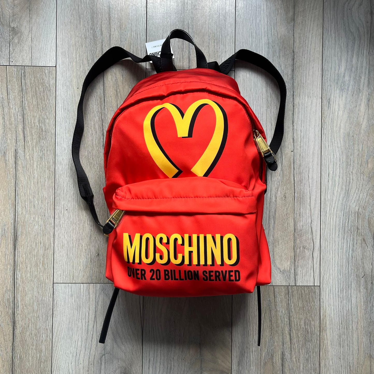 Moschino FW14 McDonald’s Fast Food Backpack is a red nylon backpack from the Autumn/Winter 2014 collection, designed by Jeremy Scott for his debut Moschino lineup. The front features yellow curved arches inspired by McDonald’s branding, with a front zip pocket printed with yellow Moschino text and black “Over 20 Billion Served” slogan. The bag includes gold-tone zippers with black leather pulls, black straps with Moschino logos, a branded interior lining, and comes with a Moschino dust bag. Made in Italy.
