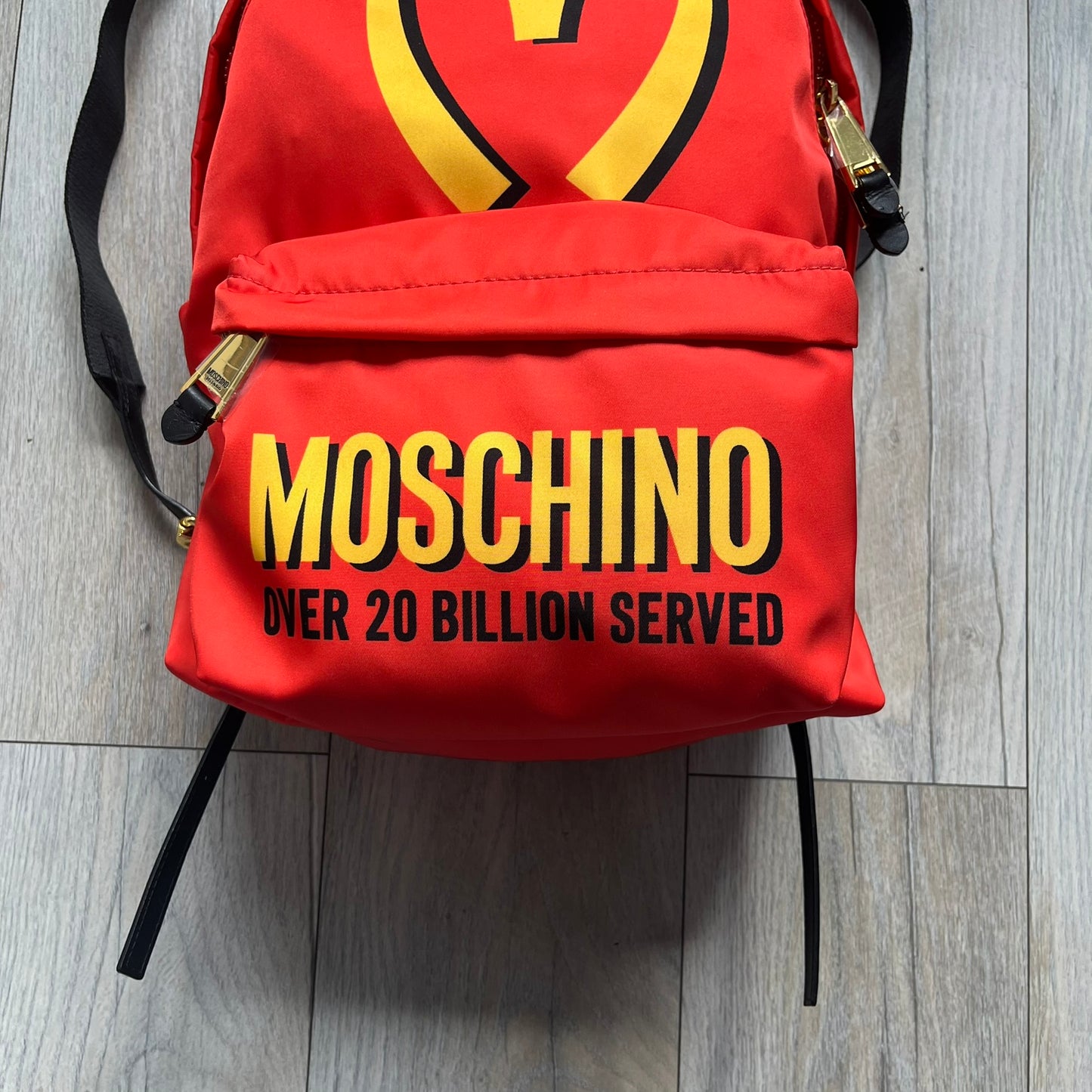 Moschino FW14 McDonald’s Fast Food Backpack is a red nylon backpack from the Autumn/Winter 2014 collection, designed by Jeremy Scott for his debut Moschino lineup. The front features yellow curved arches inspired by McDonald’s branding, with a front zip pocket printed with yellow Moschino text and black “Over 20 Billion Served” slogan. The bag includes gold-tone zippers with black leather pulls, black straps with Moschino logos, a branded interior lining, and comes with a Moschino dust bag. Made in Italy.
