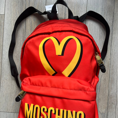 Moschino FW14 McDonald’s Fast Food Backpack is a red nylon backpack from the Autumn/Winter 2014 collection, designed by Jeremy Scott for his debut Moschino lineup. The front features yellow curved arches inspired by McDonald’s branding, with a front zip pocket printed with yellow Moschino text and black “Over 20 Billion Served” slogan. The bag includes gold-tone zippers with black leather pulls, black straps with Moschino logos, a branded interior lining, and comes with a Moschino dust bag. Made in Italy.