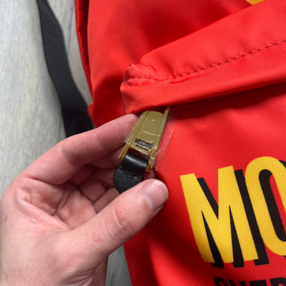 Moschino FW14 McDonald’s Fast Food Backpack is a red nylon backpack from the Autumn/Winter 2014 collection, designed by Jeremy Scott for his debut Moschino lineup. The front features yellow curved arches inspired by McDonald’s branding, with a front zip pocket printed with yellow Moschino text and black “Over 20 Billion Served” slogan. The bag includes gold-tone zippers with black leather pulls, black straps with Moschino logos, a branded interior lining, and comes with a Moschino dust bag. Made in Italy.