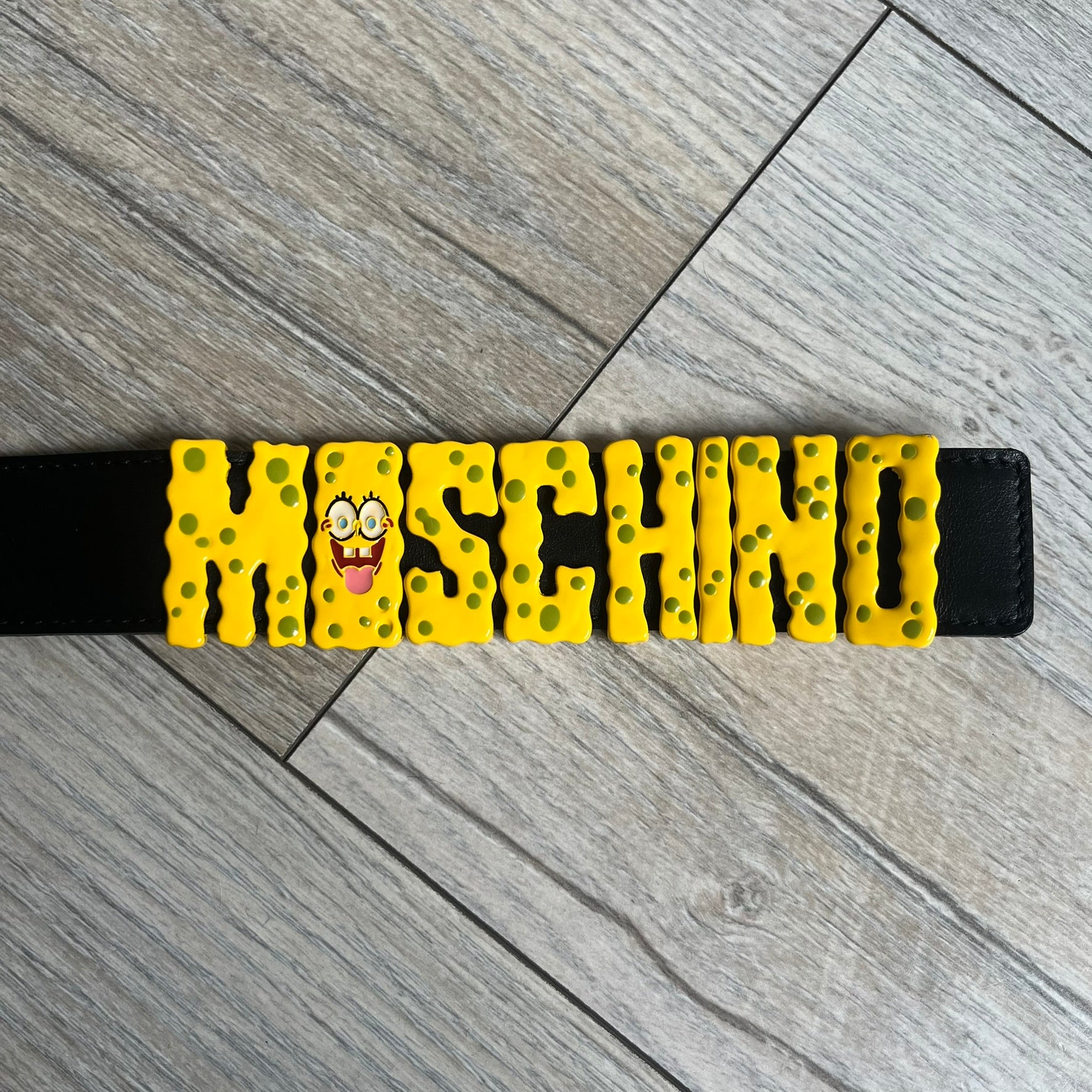 Moschino FW14 SpongeBob Leather Belt is a black leather belt from the Fall/Winter 2014 SpongeBob SquarePants collaboration. The front features the MOSCHINO logo formed by individual movable letters rather than a single rigid buckle. Each letter is finished in yellow with green sponge-like detailing, and the first “O” includes SpongeBob’s happy face. The belt uses a metal buckle closure and comes with the original dust bag and tags. Made in Italy from Genuine Leather.