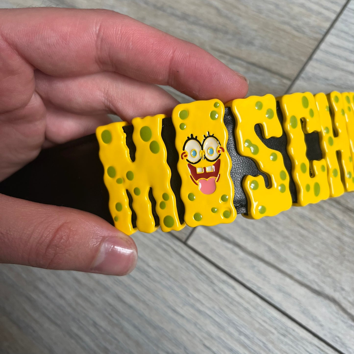 Moschino FW14 SpongeBob Leather Belt is a black leather belt from the Fall/Winter 2014 SpongeBob SquarePants collaboration. The front features the MOSCHINO logo formed by individual movable letters rather than a single rigid buckle. Each letter is finished in yellow with green sponge-like detailing, and the first “O” includes SpongeBob’s happy face. The belt uses a metal buckle closure and comes with the original dust bag and tags. Made in Italy from Genuine Leather.