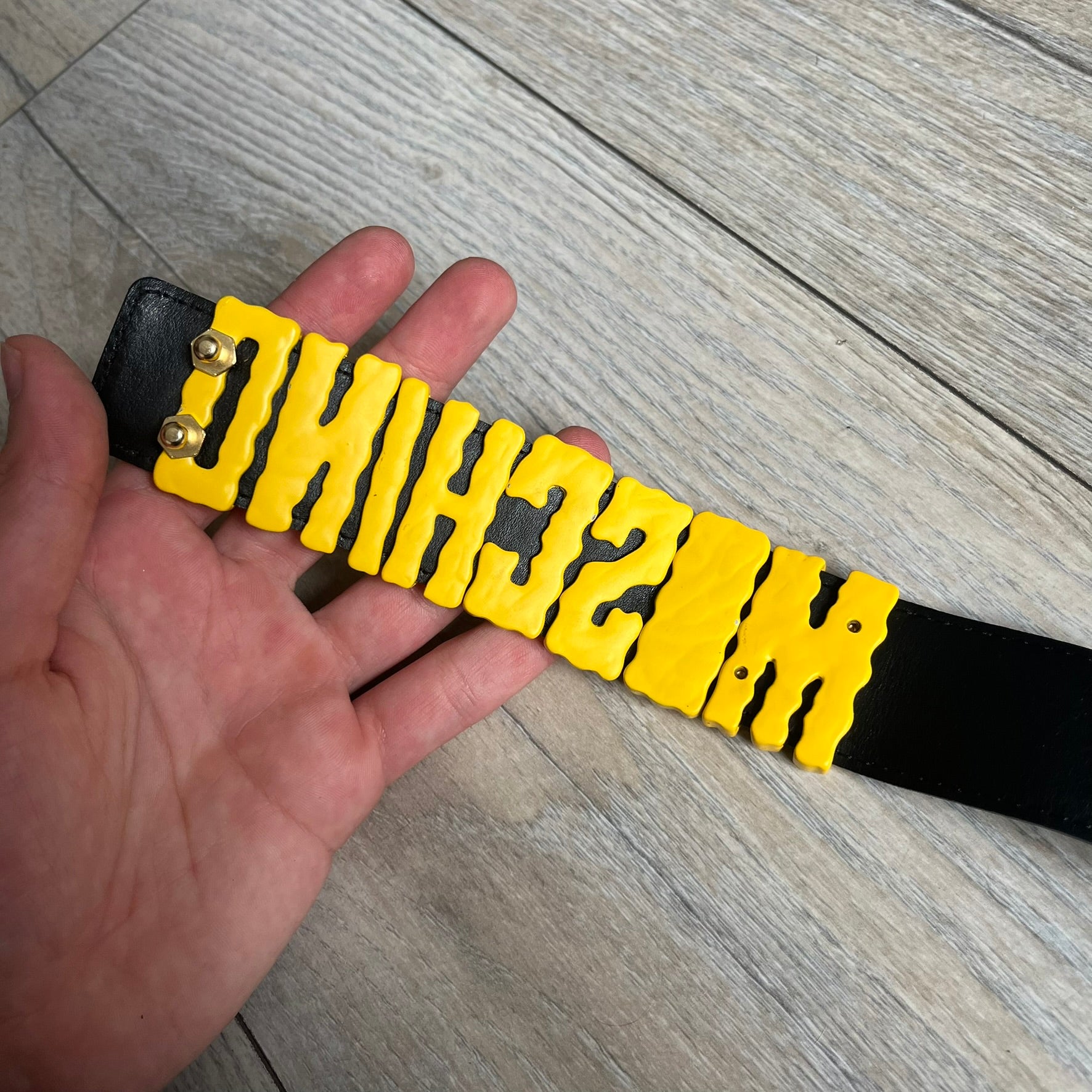 Moschino FW14 SpongeBob Leather Belt is a black leather belt from the Fall/Winter 2014 SpongeBob SquarePants collaboration. The front features the MOSCHINO logo formed by individual movable letters rather than a single rigid buckle. Each letter is finished in yellow with green sponge-like detailing, and the first “O” includes SpongeBob’s happy face. The belt uses a metal buckle closure and comes with the original dust bag and tags. Made in Italy from Genuine Leather.