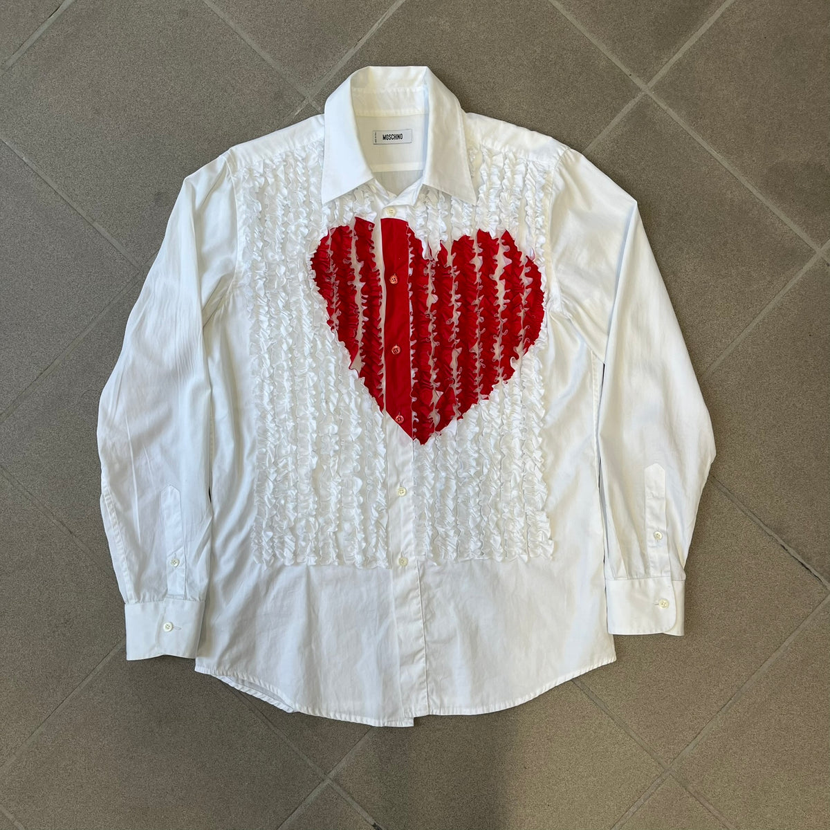 Moschino SS2000 Ruffled Heart Button Up Shirt is a white button-up made in Italy, featuring a ruffled rectangular panel on the front with an asymmetrically placed red heart at its center. It has Moschino-branded buttons, and a black Moschino text logo sits on the back just below the collar.