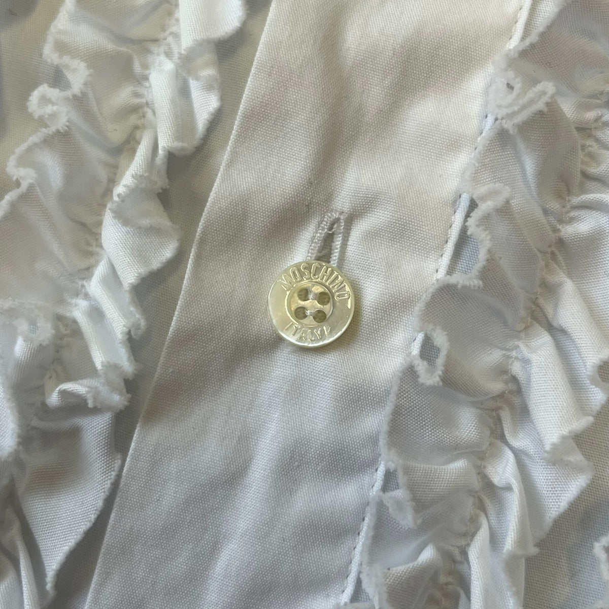 Moschino SS2000 Ruffled Heart Button Up Shirt is a white button-up made in Italy, featuring a ruffled rectangular panel on the front with an asymmetrically placed red heart at its center. It has Moschino-branded buttons, and a black Moschino text logo sits on the back just below the collar.