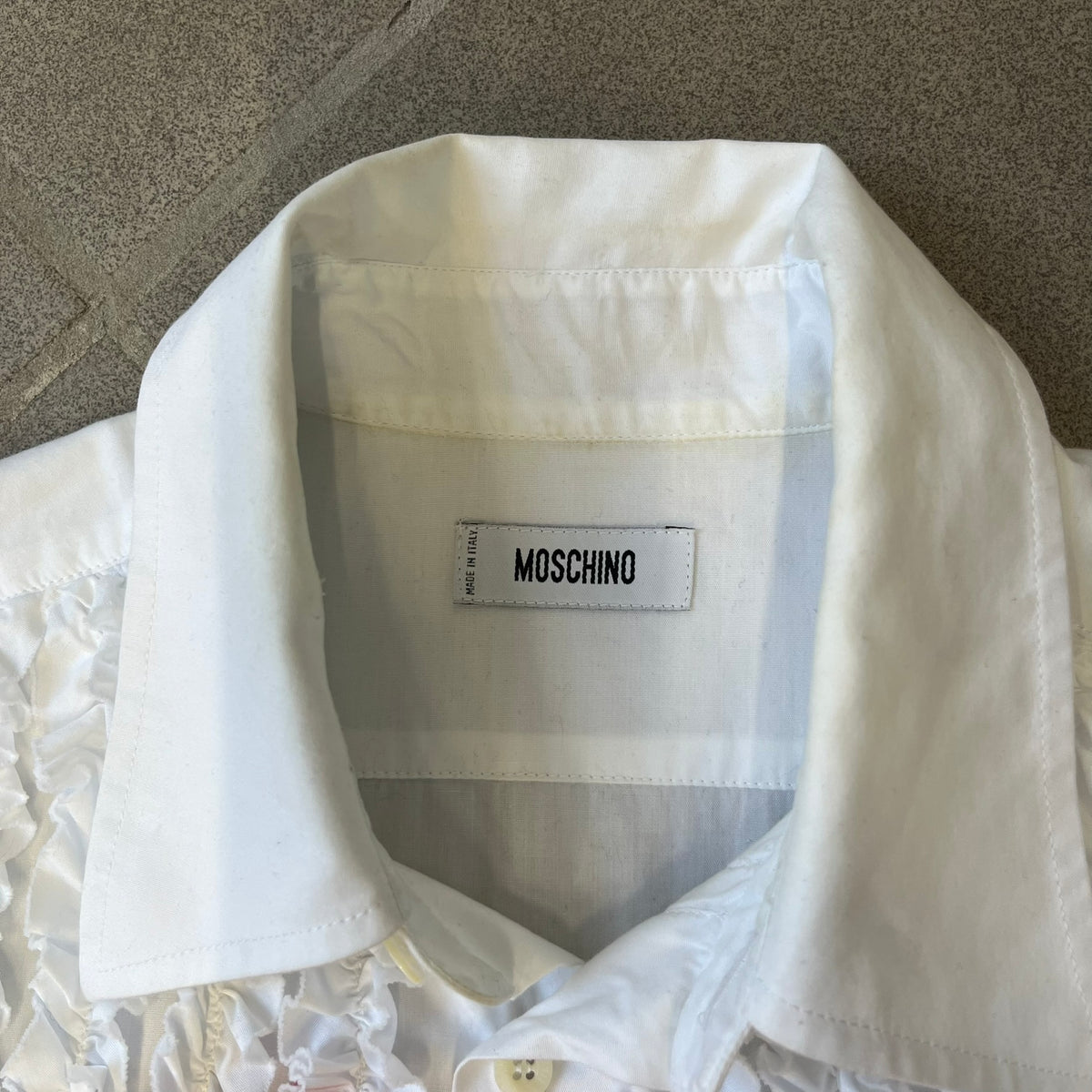 Moschino SS2000 Ruffled Heart Button Up Shirt is a white button-up made in Italy, featuring a ruffled rectangular panel on the front with an asymmetrically placed red heart at its center. It has Moschino-branded buttons, and a black Moschino text logo sits on the back just below the collar.