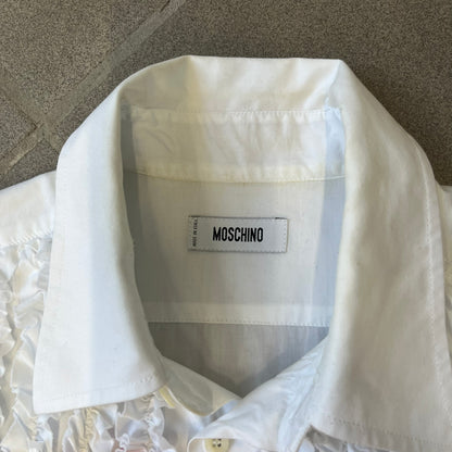 Moschino SS2000 Ruffled Heart Button Up Shirt is a white button-up made in Italy, featuring a ruffled rectangular panel on the front with an asymmetrically placed red heart at its center. It has Moschino-branded buttons, and a black Moschino text logo sits on the back just below the collar.