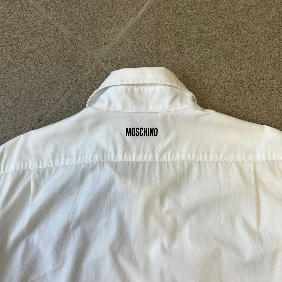Moschino SS2000 Ruffled Heart Button Up Shirt is a white button-up made in Italy, featuring a ruffled rectangular panel on the front with an asymmetrically placed red heart at its center. It has Moschino-branded buttons, and a black Moschino text logo sits on the back just below the collar.