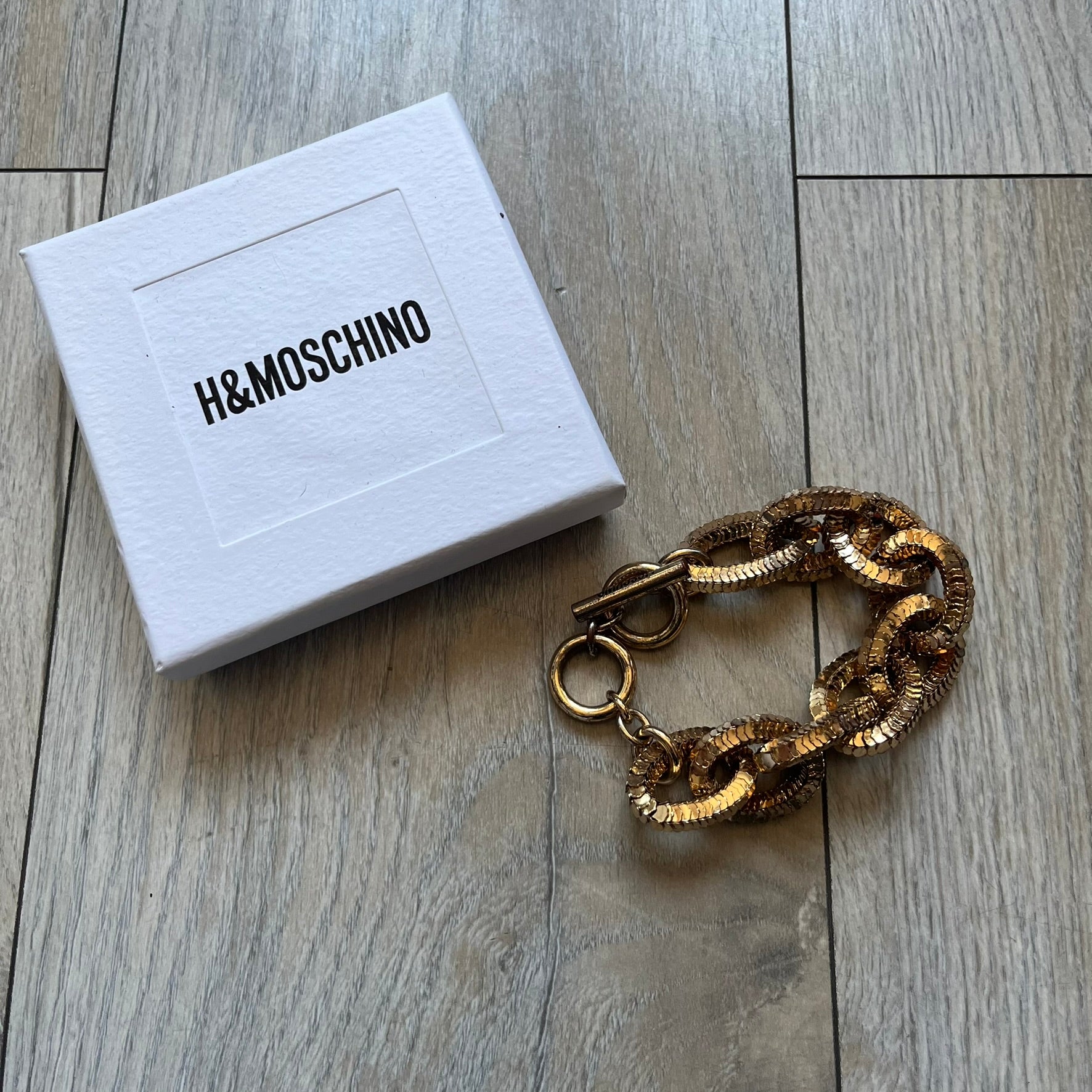 Moschino x H&M 2018 Gold Bracelet is a chunky chain bracelet released as part of the Moschino x H&M collaboration designed by Jeremy Scott. The bracelet features oversized O-shaped links in a gold-tone finish and a T-bar clasp closure. Produced for the 2018 capsule collection and made in Vietnam, it comes with the original white H&MOSCHINO box.