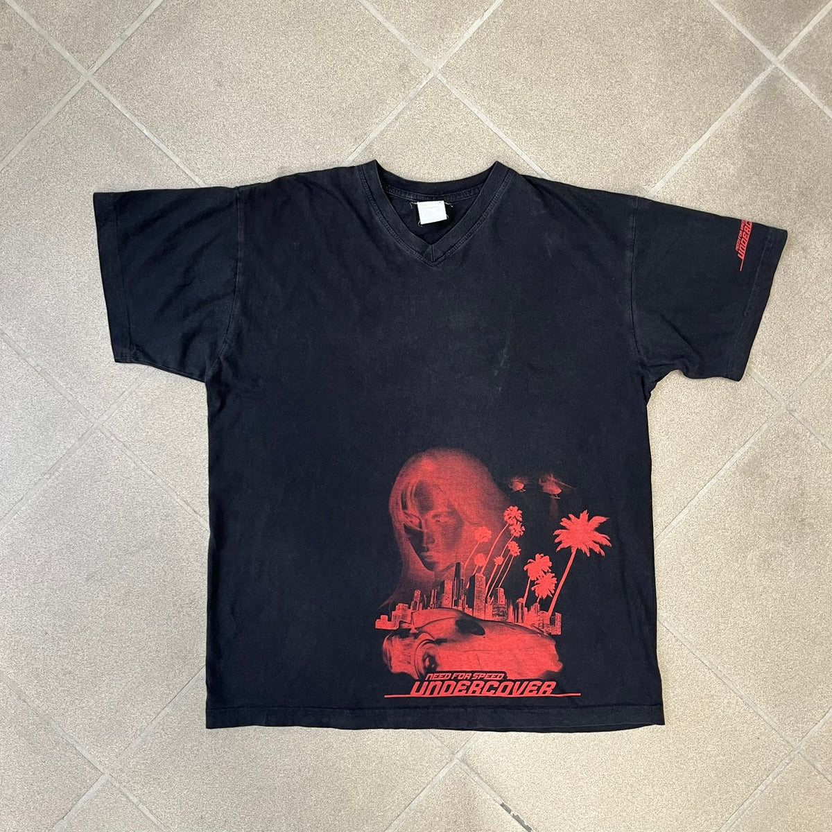 Need for Speed Undercover 2008 Promo Tee is a washed black T-shirt made on a Stedman blank, featuring a full red graphic along the bottom of a woman, palm trees, and a car with the Need for Speed Undercover logo. A red NFS logo is also printed on one sleeve.