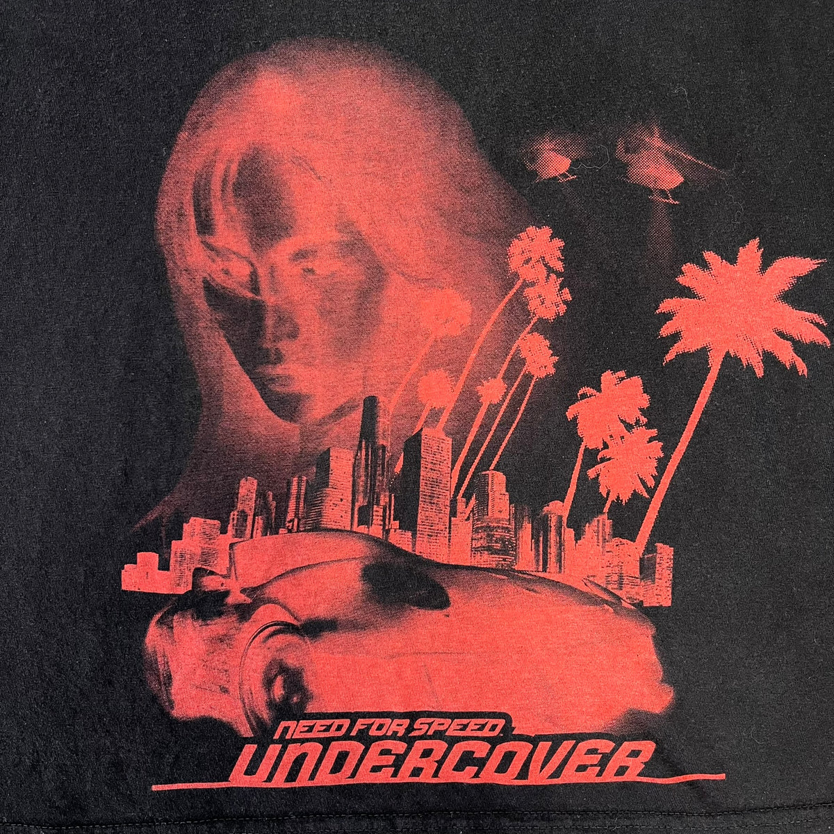 Need for Speed Undercover 2008 Promo Tee is a washed black T-shirt made on a Stedman blank, featuring a full red graphic along the bottom of a woman, palm trees, and a car with the Need for Speed Undercover logo. A red NFS logo is also printed on one sleeve.