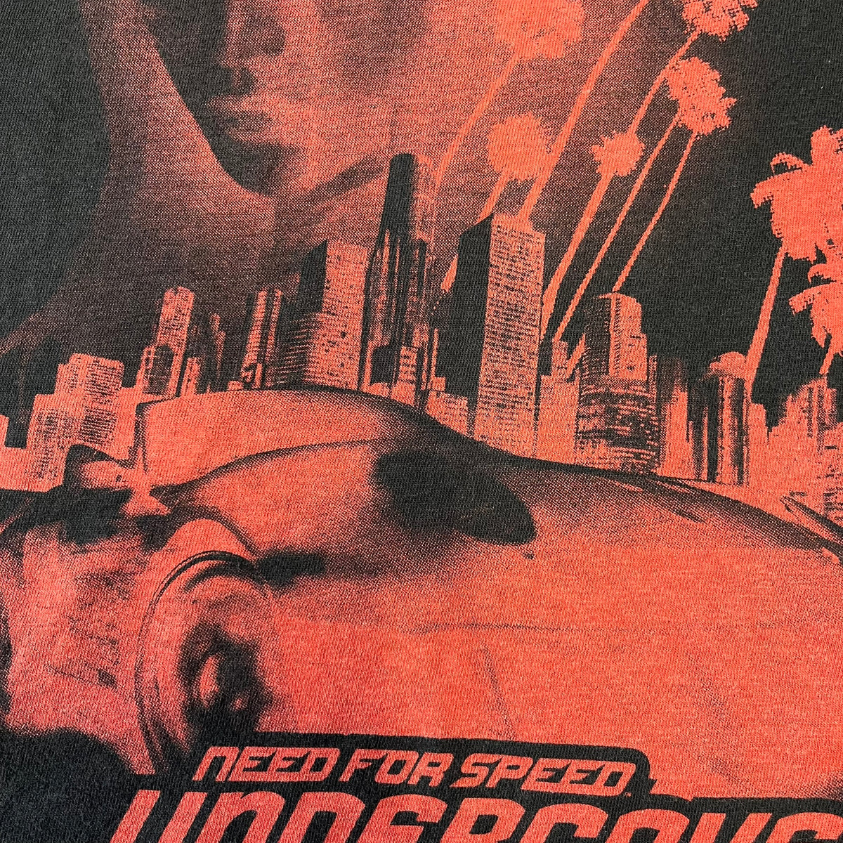 Need for Speed Undercover 2008 Promo Tee is a washed black T-shirt made on a Stedman blank, featuring a full red graphic along the bottom of a woman, palm trees, and a car with the Need for Speed Undercover logo. A red NFS logo is also printed on one sleeve.