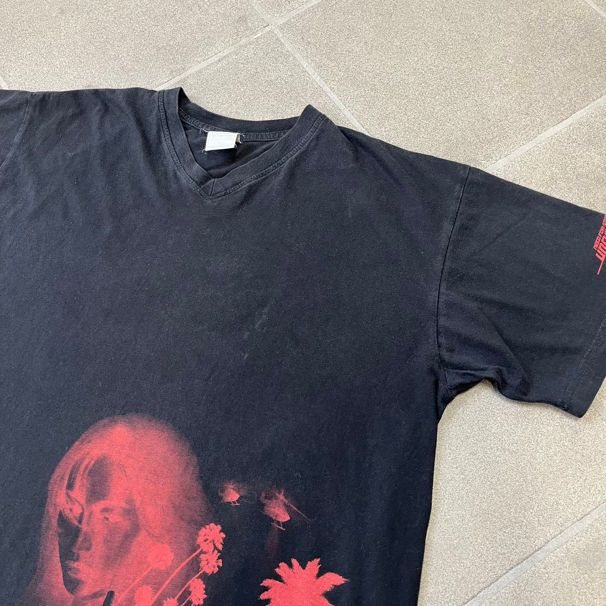 Need for Speed Undercover 2008 Promo Tee is a washed black T-shirt made on a Stedman blank, featuring a full red graphic along the bottom of a woman, palm trees, and a car with the Need for Speed Undercover logo. A red NFS logo is also printed on one sleeve.