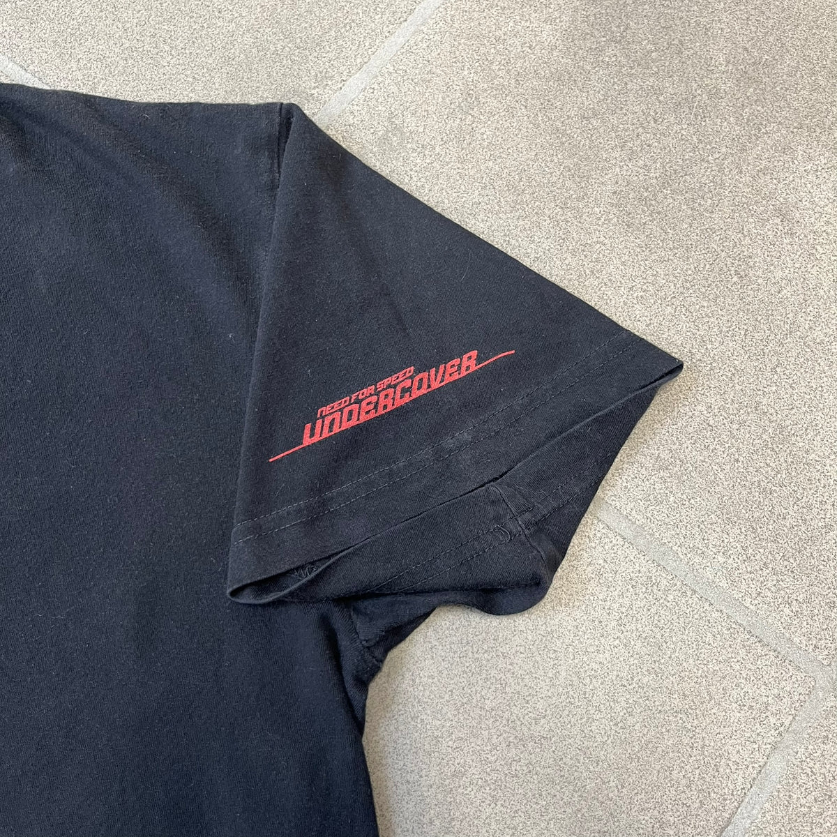 Need for Speed Undercover 2008 Promo Tee is a washed black T-shirt made on a Stedman blank, featuring a full red graphic along the bottom of a woman, palm trees, and a car with the Need for Speed Undercover logo. A red NFS logo is also printed on one sleeve.