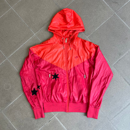 Nike 2000s Black Stars Red Windbreaker features a two-tone red design with a lighter red upper section and a darker red lower half. It is detailed with black star patches running along one sleeve and an additional star patch on the front. The jacket includes two zip pockets.