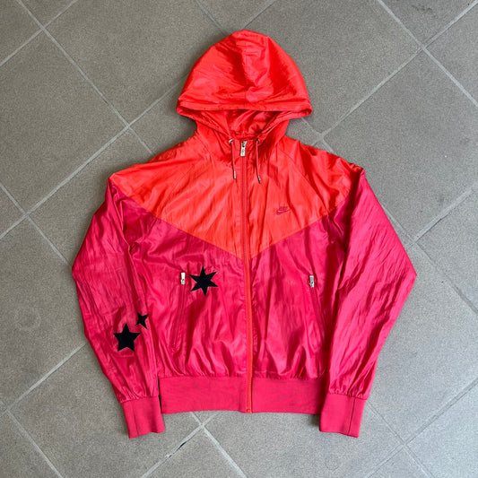 Nike 2000s Black Stars Red Windbreaker features a two-tone red design with a lighter red upper section and a darker red lower half. It is detailed with black star patches running along one sleeve and an additional star patch on the front. The jacket includes two zip pockets.