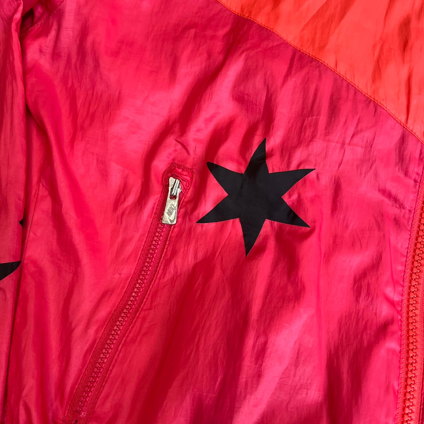 Nike 2000s Black Stars Red Windbreaker features a two-tone red design with a lighter red upper section and a darker red lower half. It is detailed with black star patches running along one sleeve and an additional star patch on the front. The jacket includes two zip pockets.