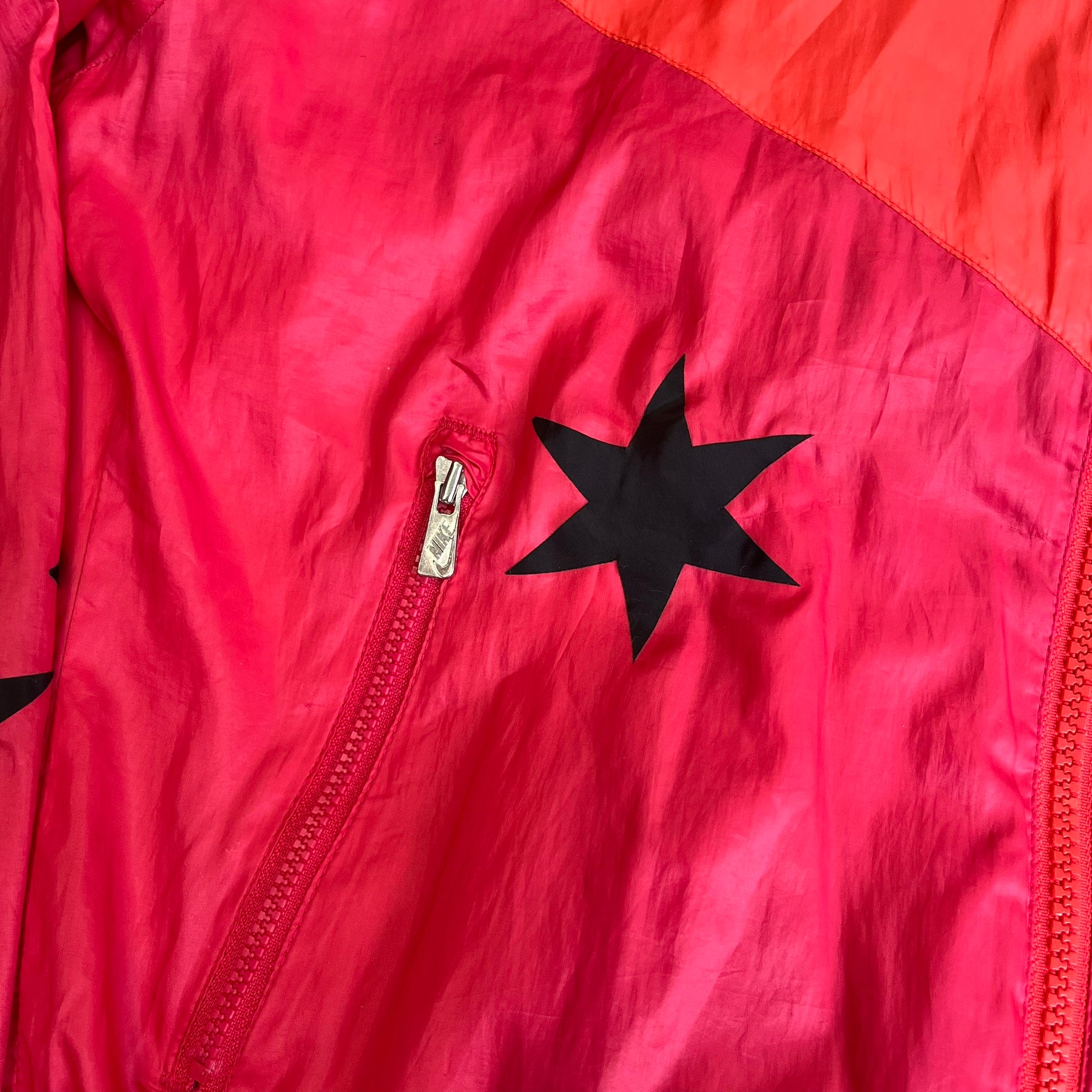 Nike 2000s Black Stars Red Windbreaker features a two-tone red design with a lighter red upper section and a darker red lower half. It is detailed with black star patches running along one sleeve and an additional star patch on the front. The jacket includes two zip pockets.