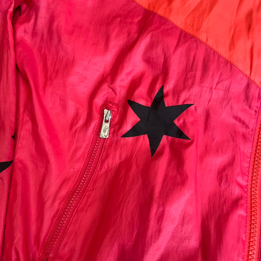 Nike 2000s Black Stars Red Windbreaker features a two-tone red design with a lighter red upper section and a darker red lower half. It is detailed with black star patches running along one sleeve and an additional star patch on the front. The jacket includes two zip pockets.