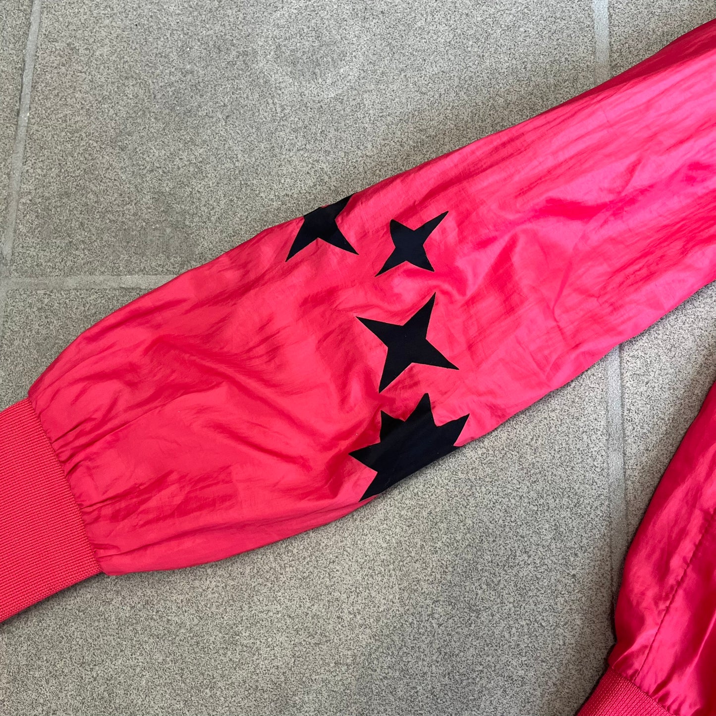 Nike 2000s Black Stars Red Windbreaker features a two-tone red design with a lighter red upper section and a darker red lower half. It is detailed with black star patches running along one sleeve and an additional star patch on the front. The jacket includes two zip pockets.