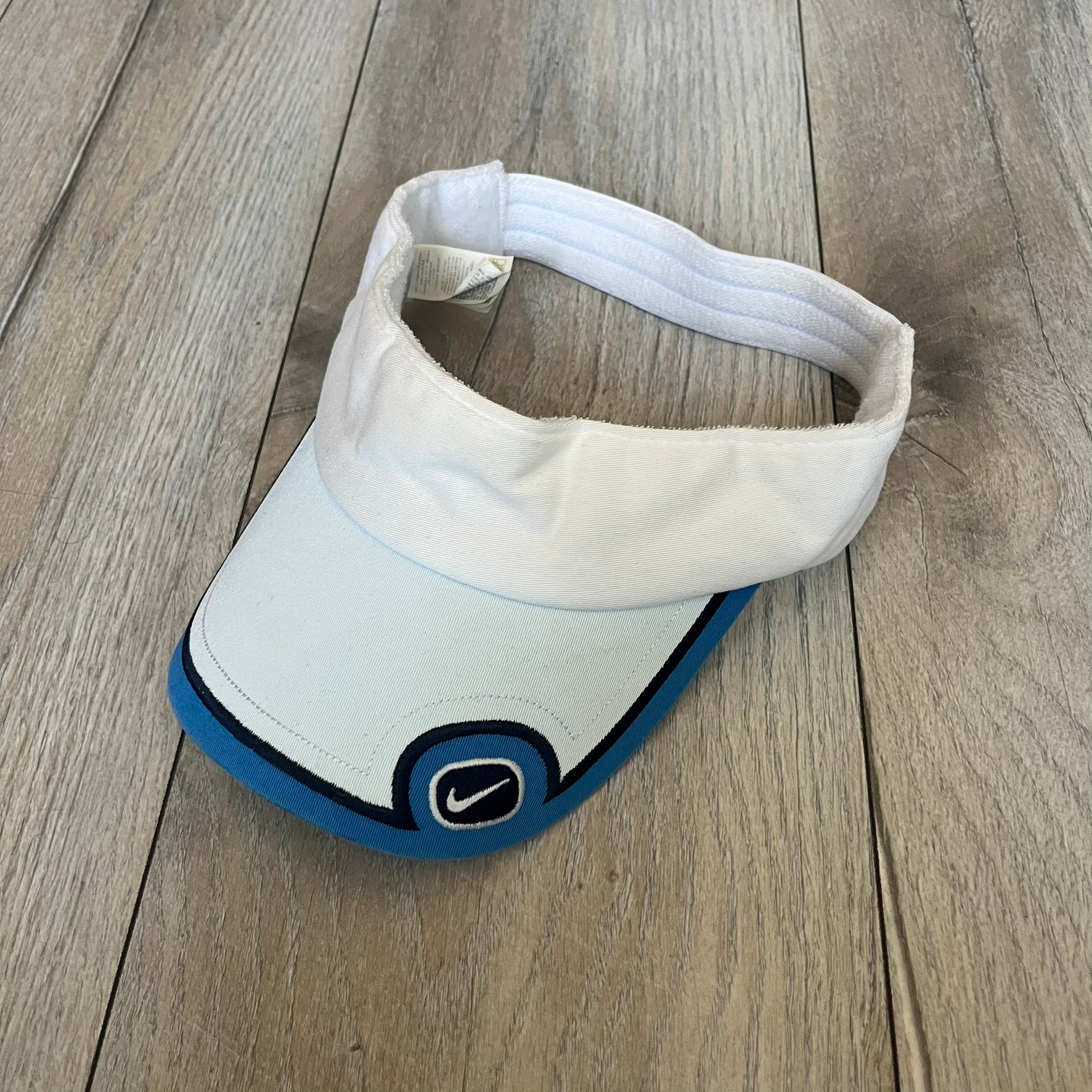 Nike 2000s Blue & White Tennis Visor is a youth-size visor cap featuring a white crown with blue trim running along the edge. The front includes a navy and white Nike logo, while the underside of the brim is finished in blue. The visor is designed with a soft, comfortable headband-style strap for a secure fit.